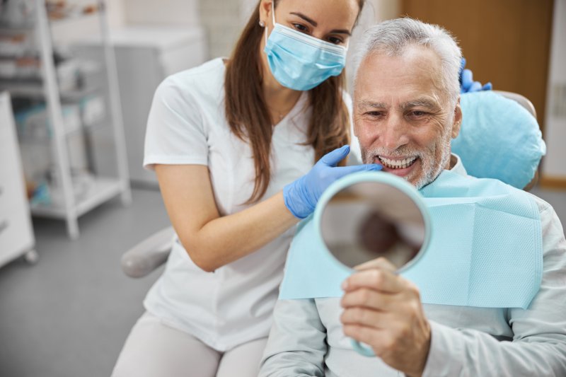 Elderly man smiling into handheld mirror as dentist wearing mask and holds gloved hand up to his chin