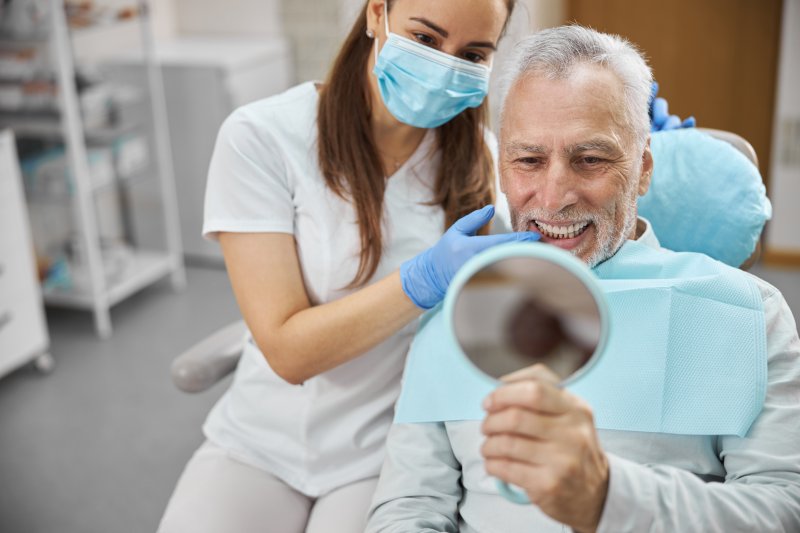 Elderly man smiling into handheld mirror as dentist wearing mask and holds gloved hand up to his chin